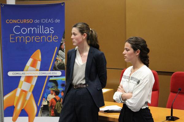 Dos mujeres están de pie frente a un banner del concurso de ideas de Comillas Emprende.