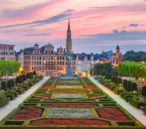 Sunset view of a formal garden with symmetrical flowerbeds leading up to historic buildings and a tall spire, under a colorful sky.