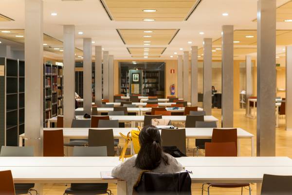 Una persona sentada en una biblioteca vacía, rodeada de mesas y estanterías.