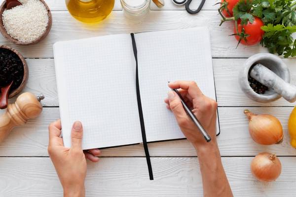 Una persona escribe en un cuaderno sobre una mesa llena de ingredientes de cocina.