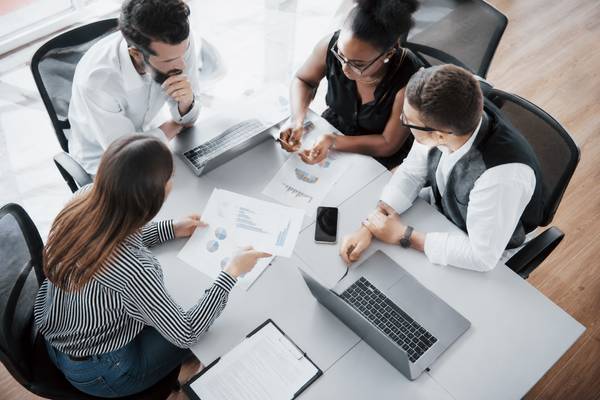 Un grupo de cuatro personas está colaborando en una reunión de trabajo con documentos y computadoras en la mesa.