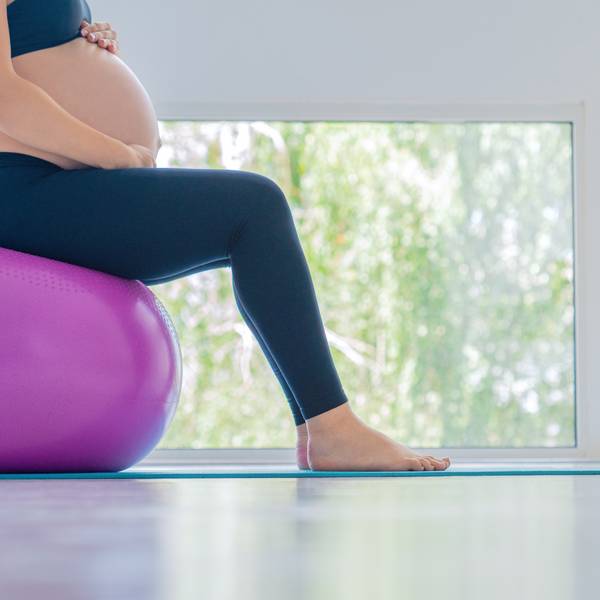 Pregnant woman sitting on a purple exercise ball near a window, practicing prenatal exercise.