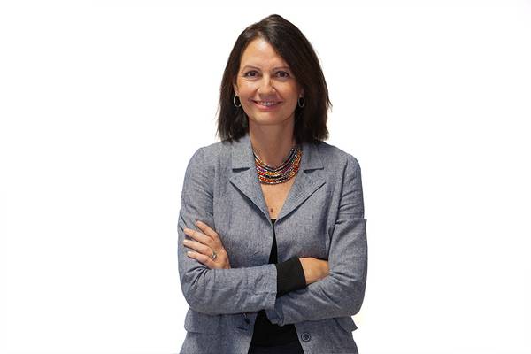 Mujer sonriente con brazos cruzados viste un traje gris y un colorido collar.