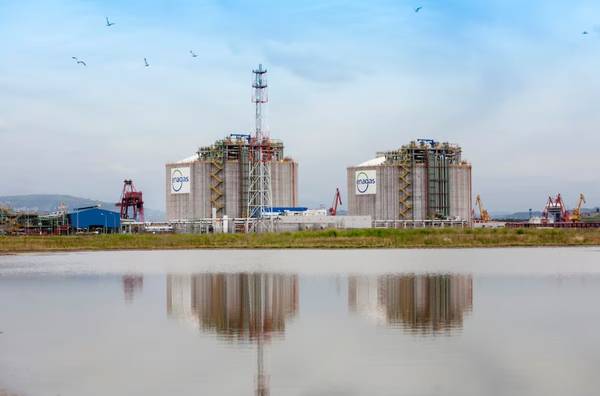 Two large industrial tanks with NASA logos reflected in a body of water, surrounded by a clear sky and flying birds.