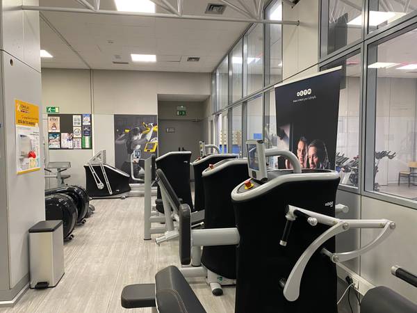 La imagen muestra un gimnasio moderno con diversas máquinas de ejercicio dispuestas en un ambiente luminoso.