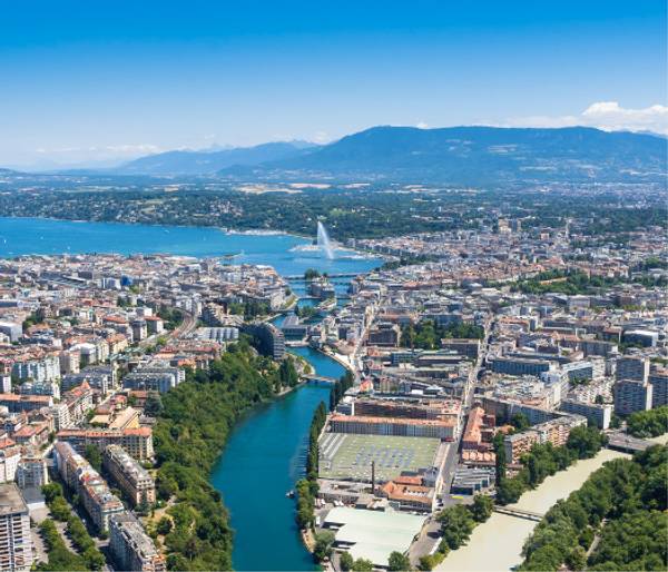 Aerial view of Geneva, showcasing the cityscape with its distinctive water jet, surrounded by the river, lake, and mountains in the background.