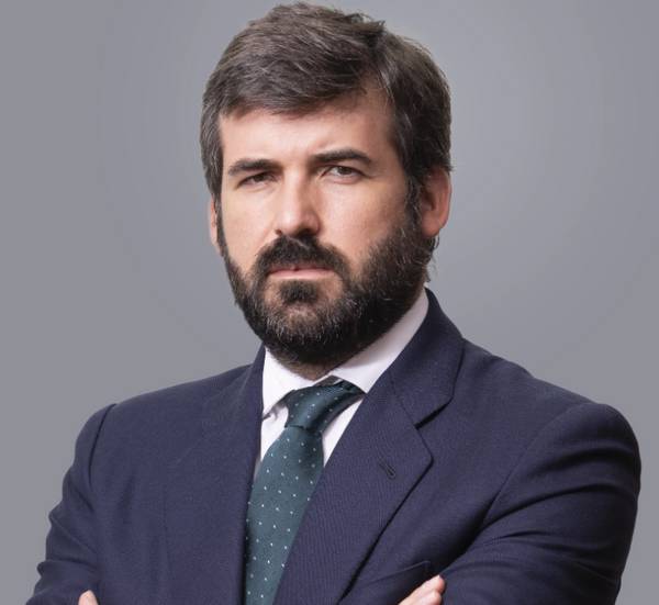 Retrato de un hombre con barba, vestido formalmente en un ambiente neutro.