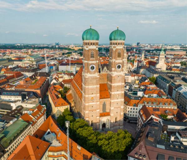 Aerial view of a large cathedral with twin green domes surrounded by dense urban buildings under a clear sky.