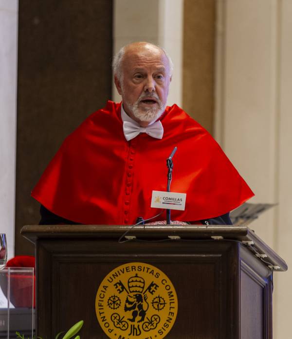 Un hombre con barba y toga roja dando un discurso desde un podio que lleva el emblema de la Universidad Pontificia Comillas.