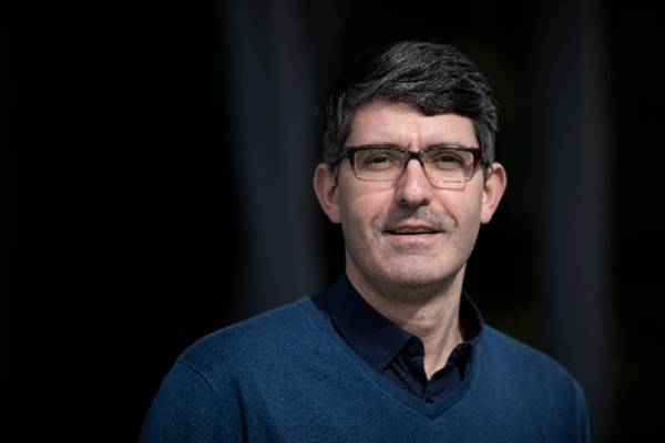 A portrait of a middle-aged man with glasses, wearing a blue sweater over a collared shirt, standing outdoors.