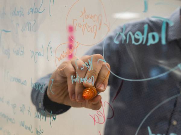 Una persona escribe en una pizarra con un marcador naranja mientras se ven anotaciones en distintas colores.