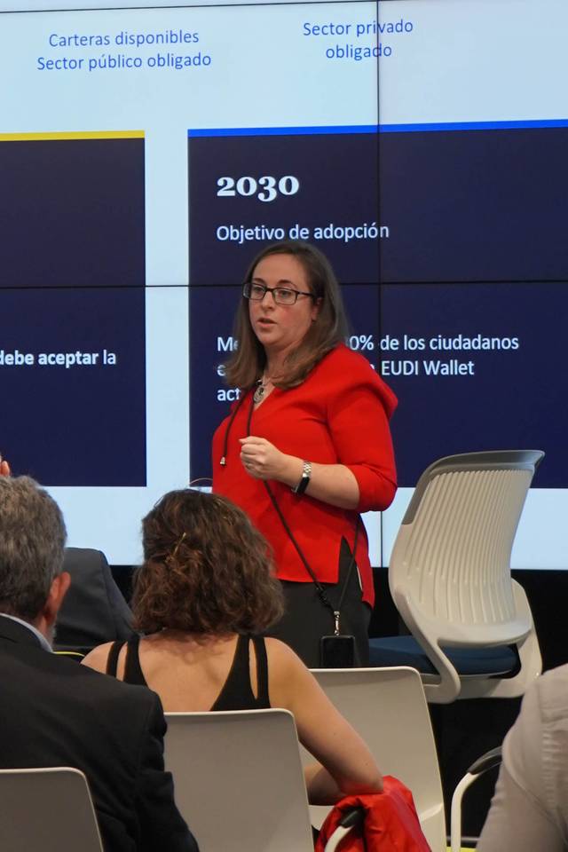 Una mujer de cabello largo y gafas, vestida con una chaqueta roja, presenta información en una sala con pantallas digitales en el fondo.