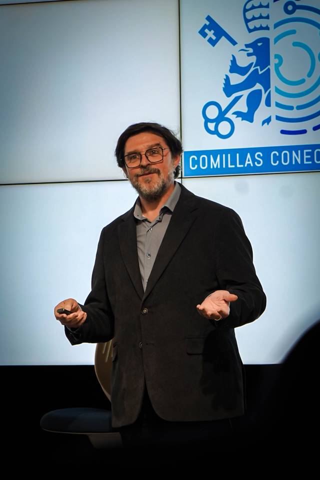 Un hombre con gafas y chaqueta oscura presenta en un evento con una pantalla detrás.