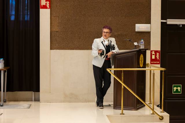Una mujer de pie junto a un podio en un espacio interior, vestida con un traje blanco y negro, mientras sostiene un dispositivo en la mano.