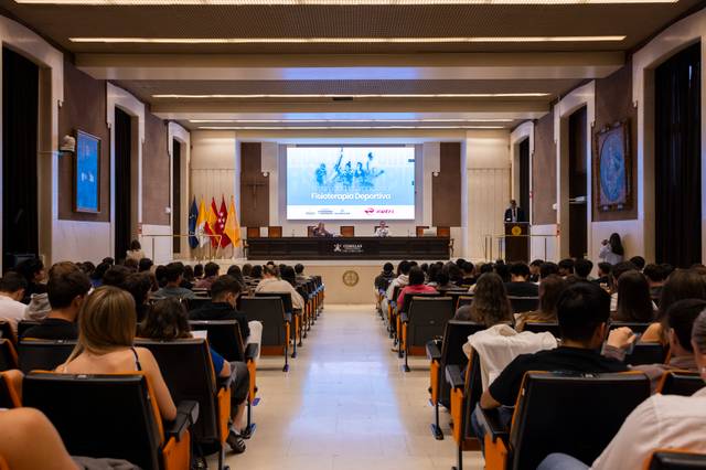 Una conferencia en un auditorio con numerosos asistentes atentos frente a un escenario.