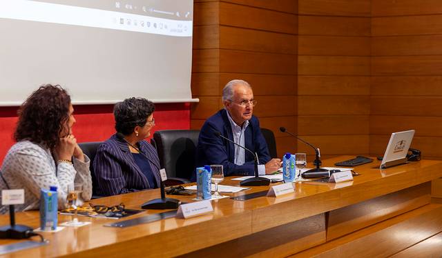 Three people sitting at a conference table during a presentation in a lecture hall.