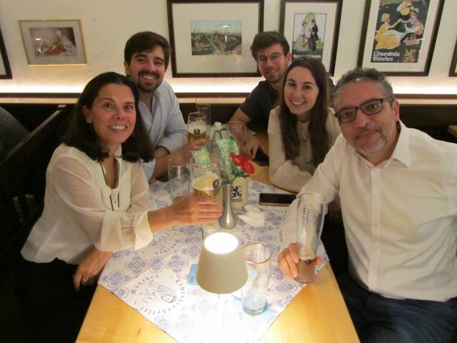 A group of six people smiling and sitting around a table in a restaurant, holding drinks.