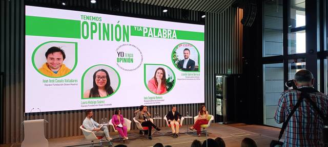 A photograph of a panel discussion event with participants seated on stage below a large screen displaying their names and photos.