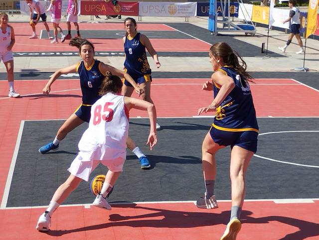 Women playing a basketball game outdoors on a red court.