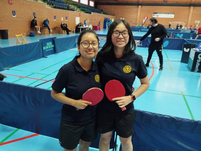 Two women standing indoors holding table tennis paddles with a sports setting in the background.