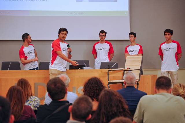 A group of five young men in matching red and white t-shirts are presenting in front of an audience at a university lecture hall.