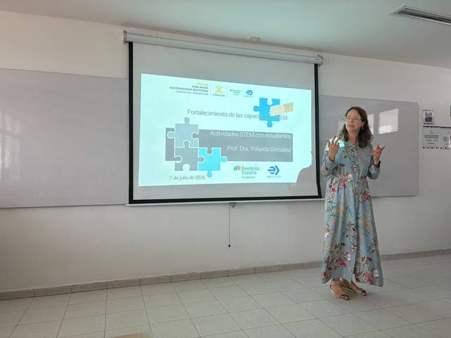 Una mujer presenta un proyecto educativo en un aula con una pantalla detrás que muestra un título sobre el fortalecimiento de capacidades en STEM.