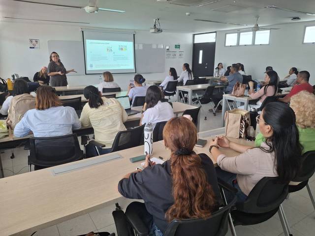 Una conferencia se lleva a cabo en un aula con varios asistentes sentados mientras una persona presenta frente a la pantalla.