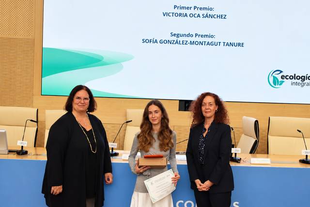 La imagen muestra a tres mujeres en una ceremonia de premiación, con dos de ellas sosteniendo diplomas y el escenario de fondo indicando los premios.