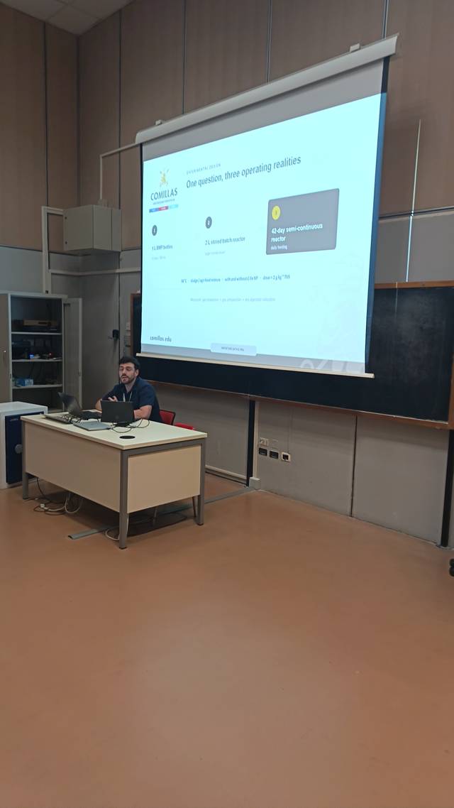 Un hombre presenta un tema frente a una pantalla en un aula.