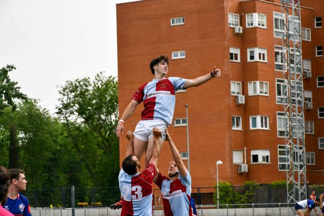 Jugadores de rugby elevando a un compañero durante un lineout en un campo al aire libre con un edificio de apartamentos al fondo.