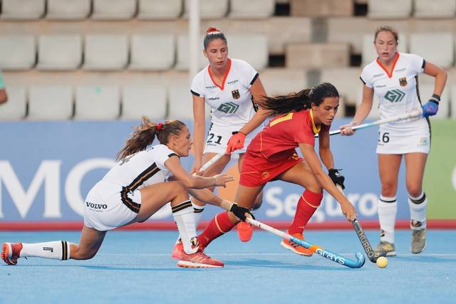 Una jugadora de hockey sobre césped vistiendo camiseta roja disputa el control del balón con una adversaria de camiseta blanca