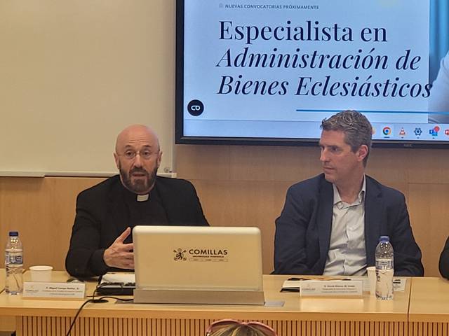 A clergyman and a man in a suit are seated at a table during a presentation about 'Especialista en Administración de Bienes Eclesiásticos' at Comillas University.