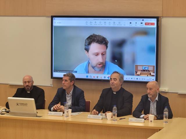 A group of men sitting at a conference table with a large screen displaying a video call in the background.
