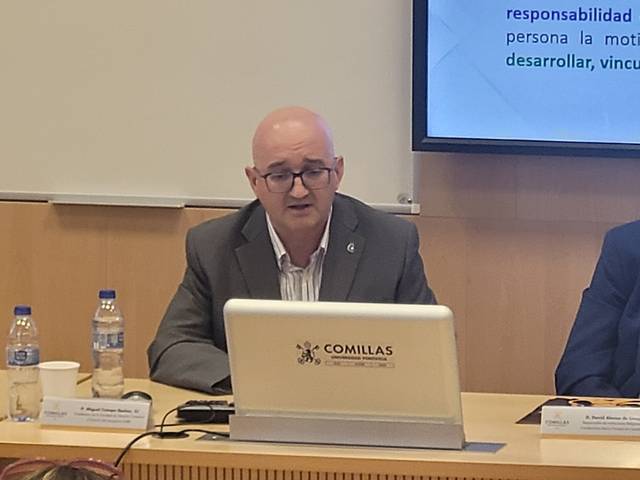 A man sitting at a desk with a laptop in a conference room, presenting at an event labelled 'Comillas'.