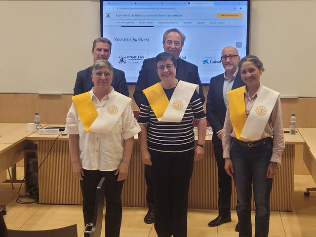 A group of six people, three of them wearing sashes, standing in a seminar room with a large screen displaying a webpage in the background.