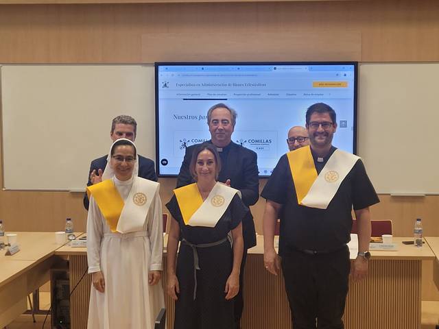 A group of five people smiling in a conference room, two wearing ceremonial sashes, standing in front of a presentation screen.