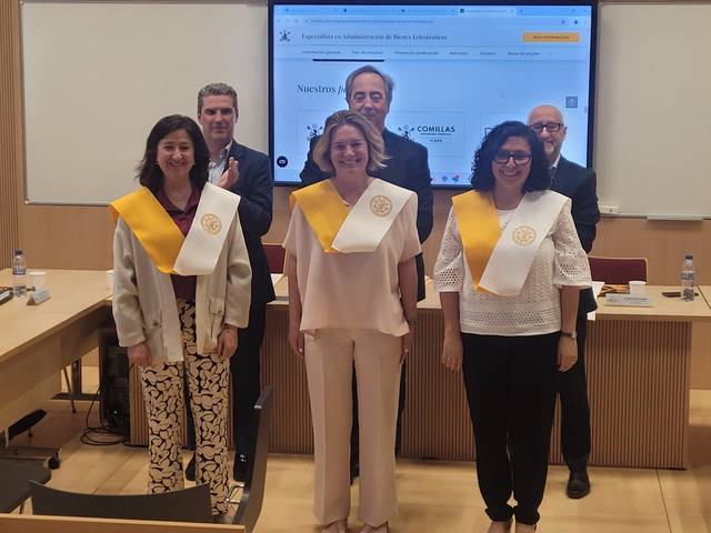 Five people wearing academic sashes standing in a classroom, smiling at a ceremony.