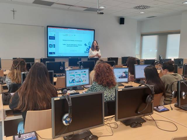 Una mujer impartiendo una clase en un aula de informática con varios estudiantes atentos a su presentación.