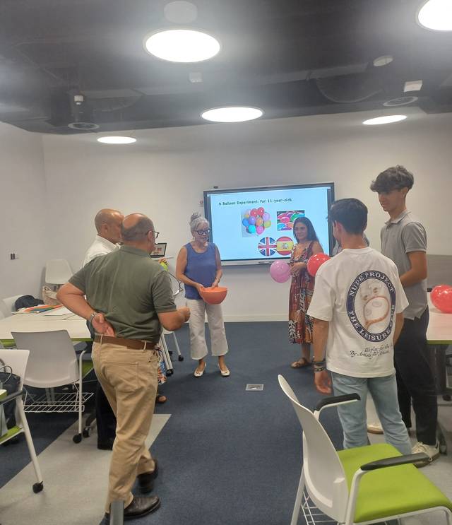 Un grupo de personas en un aula participan en una actividad con globos y una presentación en pantalla.