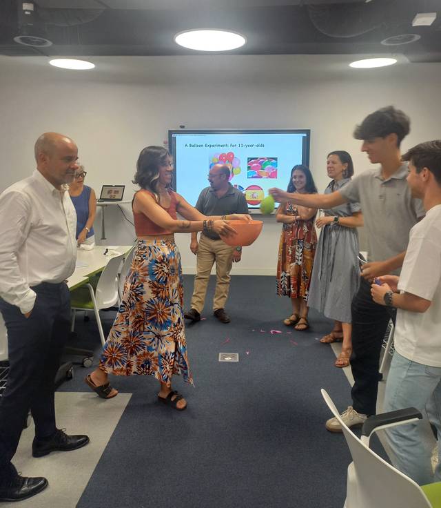 Un grupo de personas participan en una actividad en una sala de reuniones, donde una mujer sostiene un recipiente mientras otros le entregan una pelota.