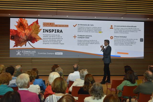 Una presentación en un auditorio, donde un ponente habla frente a una multitud mientras una presentación visual está proyectada en la pantalla.