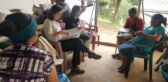 Un grupo de personas de diversas edades sentadas alrededor de mesas en un ambiente semiabierto, participando en una actividad de escritura o lectura.