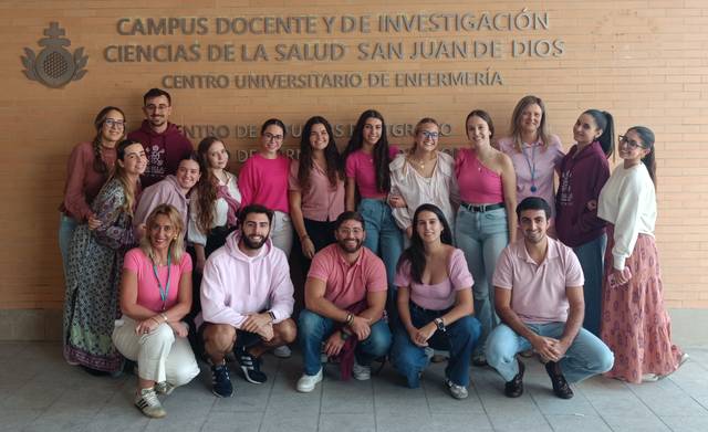 Grupo de personas posando en un campus universitario, todos vestidos con tonos de rosa frente a un mural institucional.