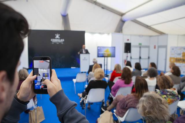 Un hombre grabando a un ponente que se encuentra en un escenario frente a una audiencia en un evento.