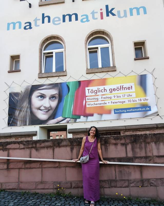Una mujer de pie frente a un edificio con un gran cartel que dice 'Täglich geöffnet' en alemán.