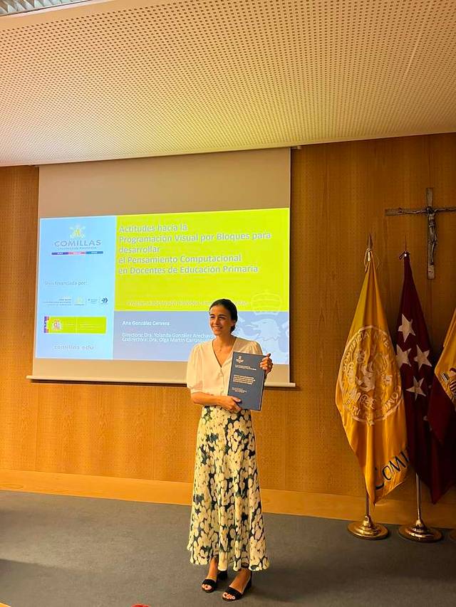 Una mujer sonriente sostiene un documento en un evento académico en una sala con banderas y una presentación en la pared.