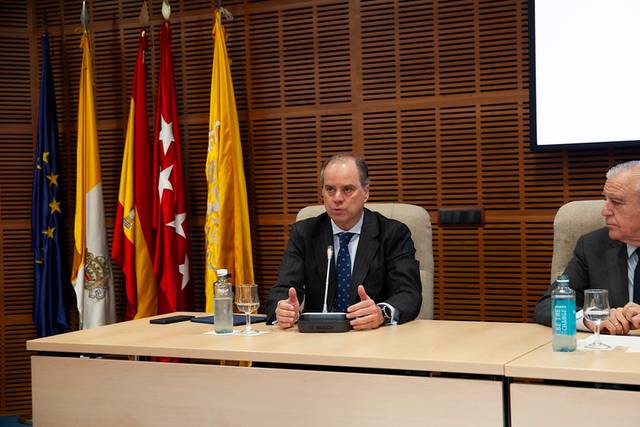 A man in a suit is speaking at a conference table with microphones and flags in the background.