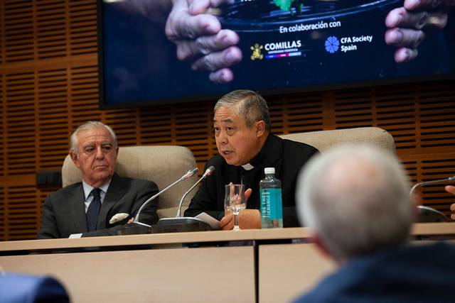 Two men, one speaking, are seated at a panel discussion in a conference room with an audience in front.