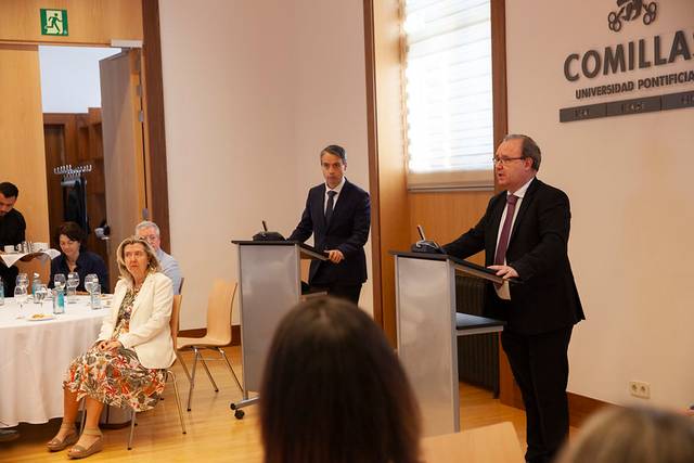 Two men are speaking at podiums in a conference room at Comillas Pontifical University, with an audience and a seated woman in the foreground.