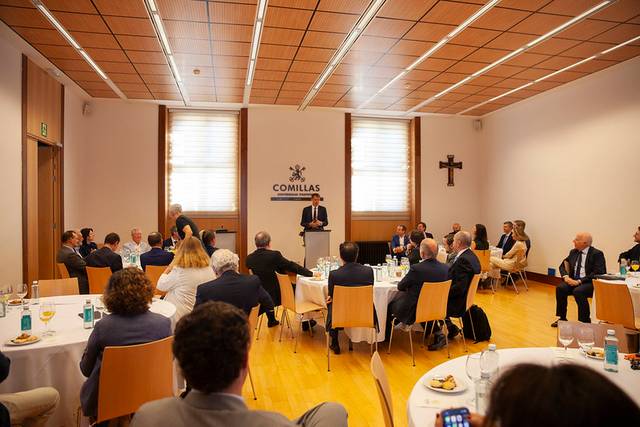 A speaker is addressing an audience in a formal indoor gathering at a venue with the logo 'COMILLAS' in the background.
